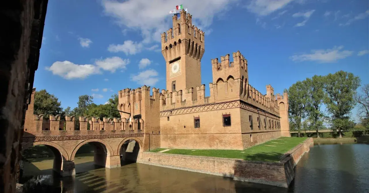 Un imponente castello in mattoni rossi, uno dei tanti castelli Emilia Romagna, con un ponte ad archi sull'acqua e torri merlate sotto un cielo azzurro.