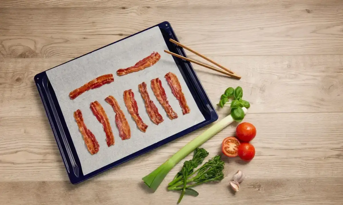 Bacon i ugn på bakplåtspapper, bredvid grönsaker och vitlök. Perfekt för en god måltid.