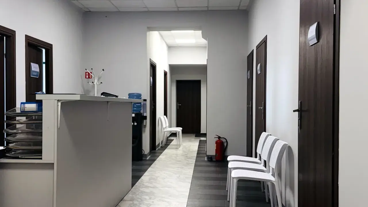 Korytarz kliniki medycznej z recepcją, drzwiami i białymi krzesłami.
