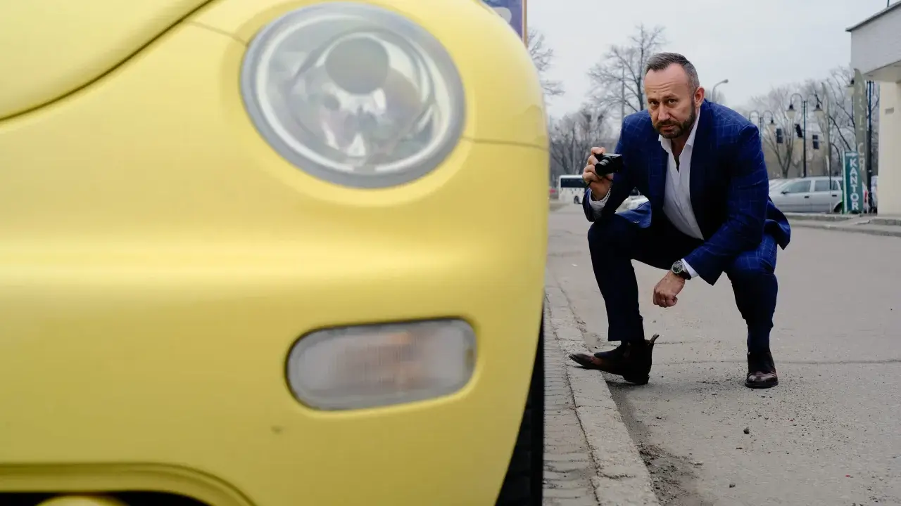 Rzeczoznawca samochodowy w garniturze klęczy przy żółtym samochodzie, robiąc zdjęcie aparatem.
