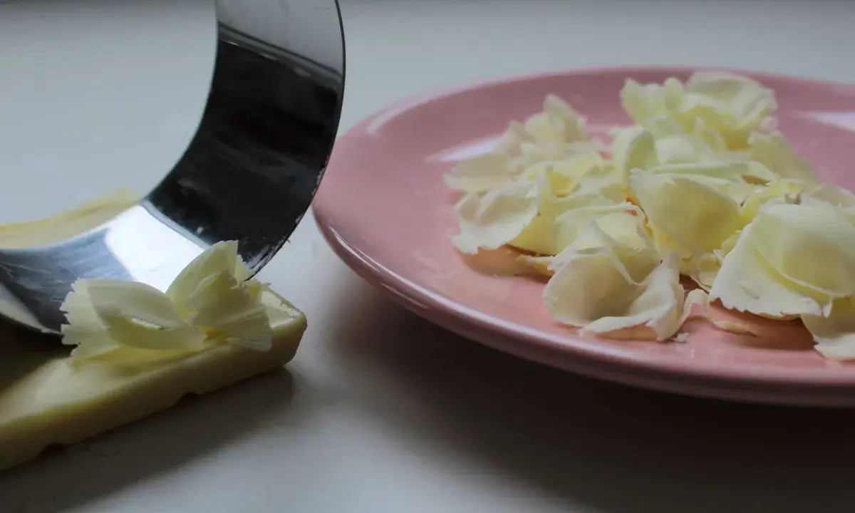 Un ustesile en métal crée des copeaux de chocolat blanc sur une assiette rose.