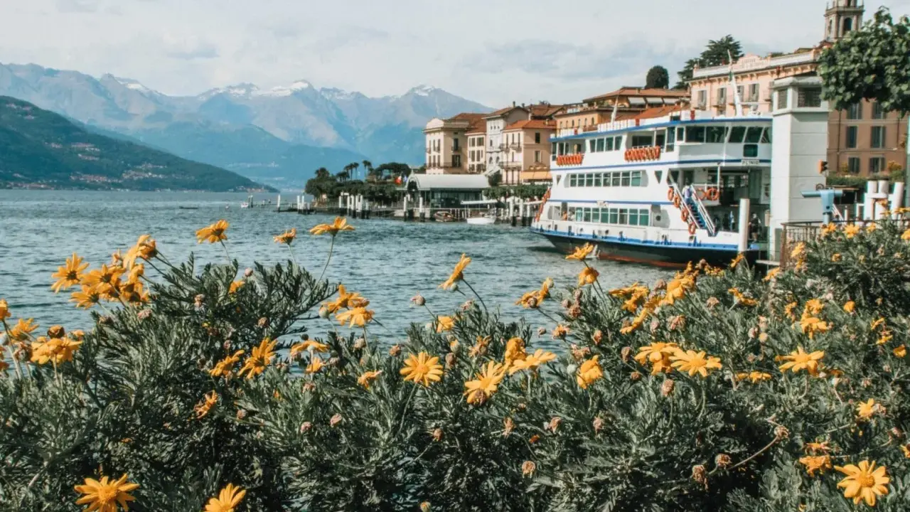 Fiori gialli in primo piano, lago blu, montagne innevate e un battello attraccato. Una perfetta gita fuori porta Milano.