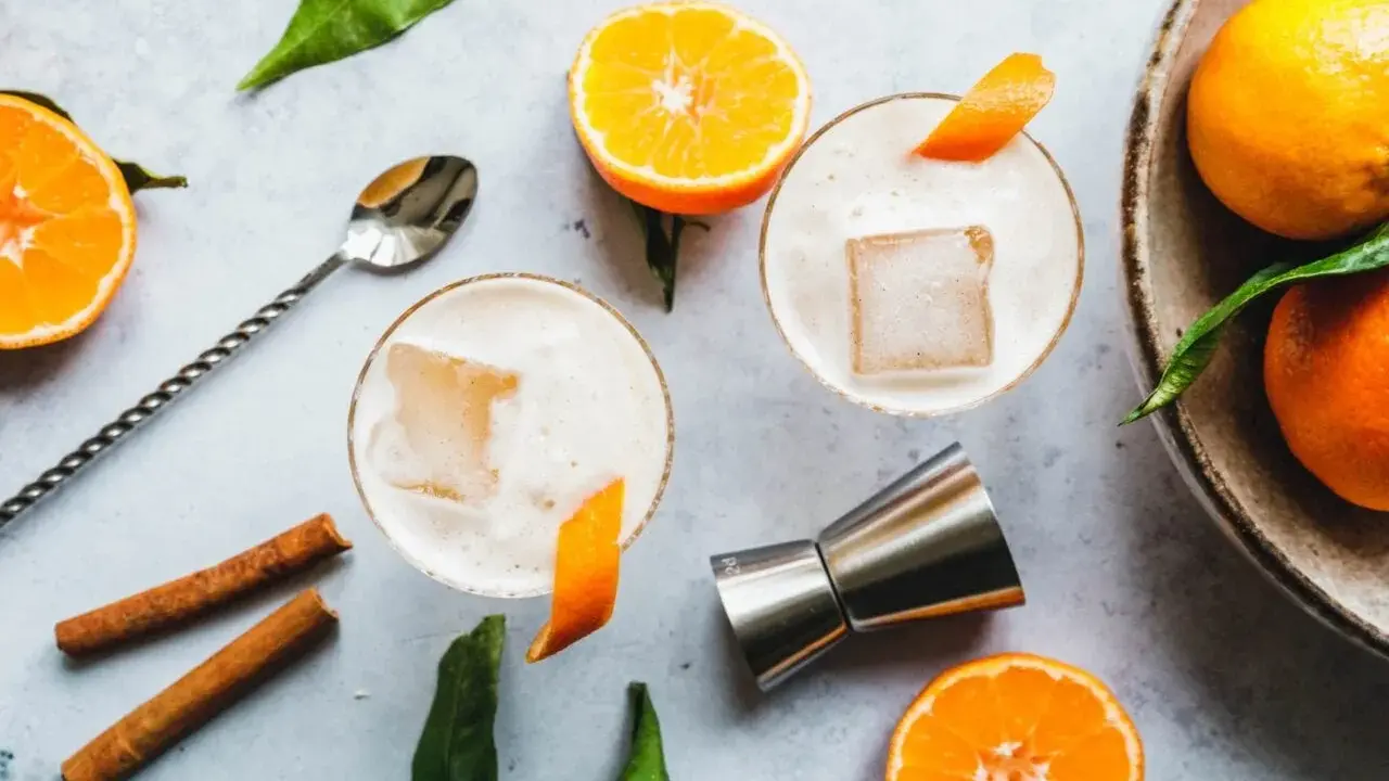 Zwei cremige Gin Sours mit Eis und Orangenzesten, garniert mit Zimtstangen und frischen Mandarinen.
