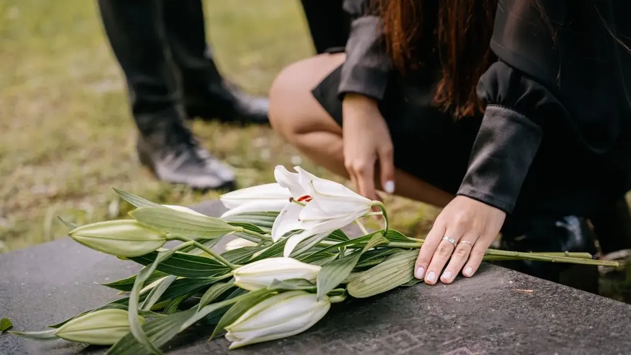 Dłoń kładzie białe lilie na kamieniu nagrobnym. W tle widać nogi w czarnych butach. To symboliczny gest, który może nastąpić w dowolnym momencie po śmierci, nie ma ustalonego terminu, ile dni po śmierci jest pogrzeb.