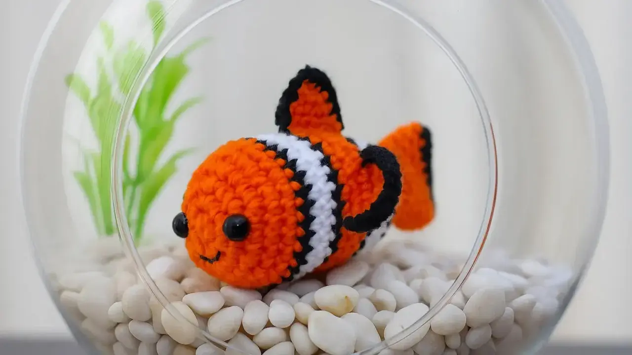 Adorable pez payaso de ganchillo en una pecera con piedras blancas y plantas verdes. ¡Perfecto para patrones de peces de ganchillo!