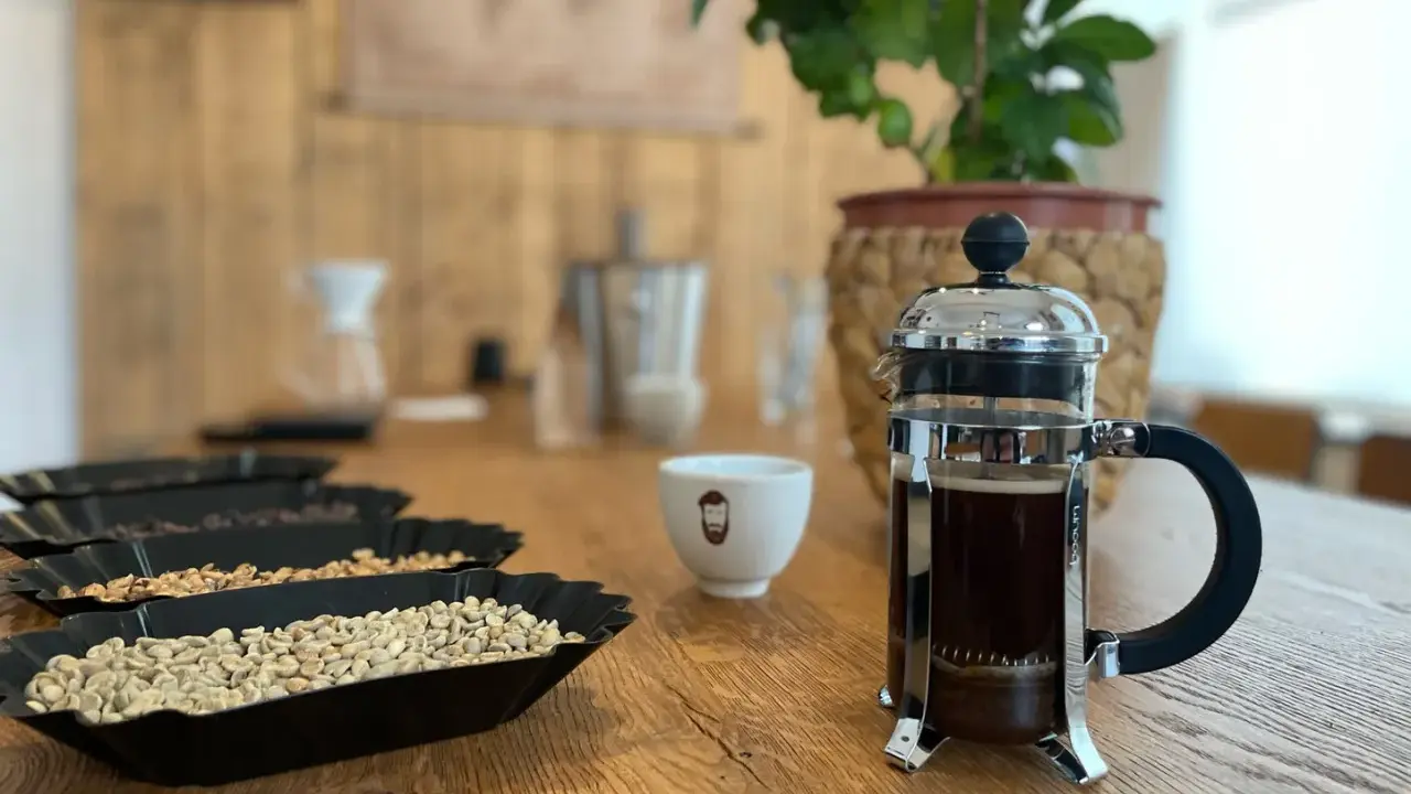 Eine French Press mit frisch gebrühtem Kaffee steht neben einer Tasse und Schalen mit Kaffeebohnen.