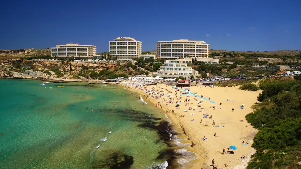 En livlig strand med turkost vatten och gyllene sand vid Golden Bay, Malta. Flera hotell syns i bakgrunden.