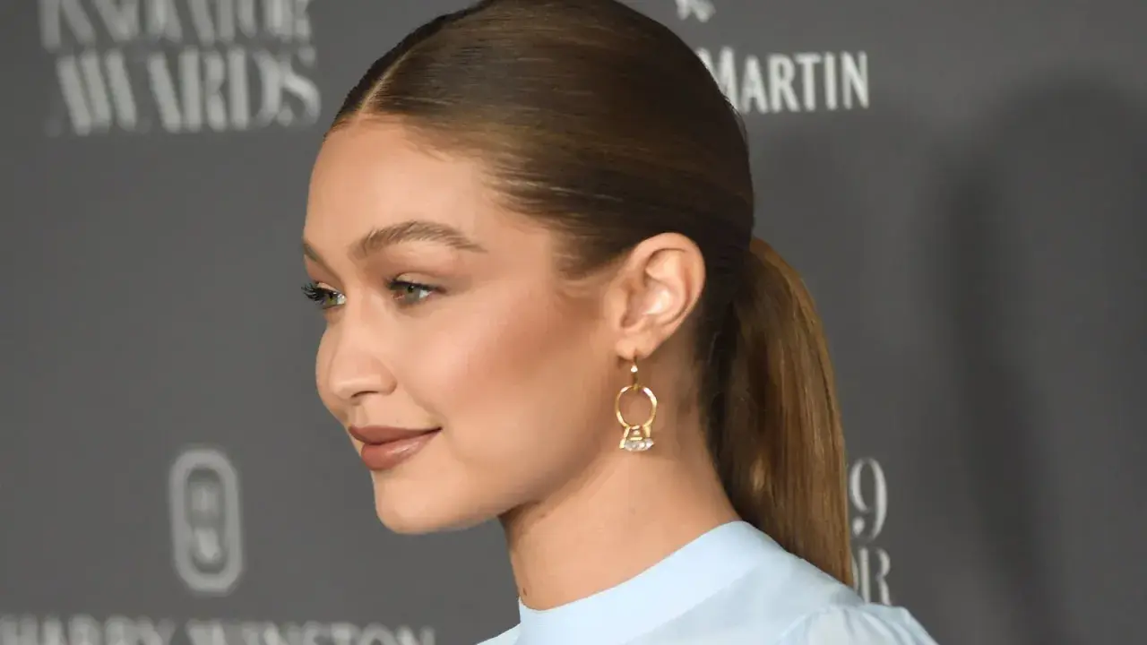 Gigi Hadid mit glatten, **haare nach hinten** gebundenen Haaren und einem dezenten Lächeln. Sie trägt ein hellblaues Oberteil und auffällige Ohrringe.
