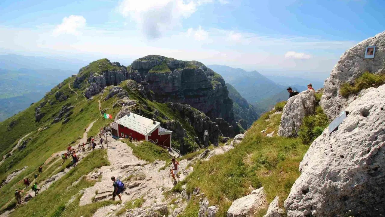 Sentiero panoramico verso il rifugio Lecco, con escursionisti su un crinale erboso e roccioso sotto un cielo azzurro.