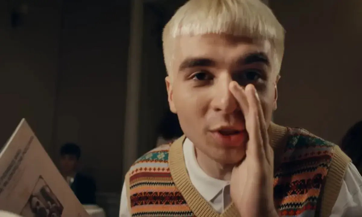 Młody mężczyzna z blond fryzurą, w kamizelce z kolorowym wzorem, trzyma rękę przy ustach, jakby coś szeptał. Obok niego leży blok mata tekst.