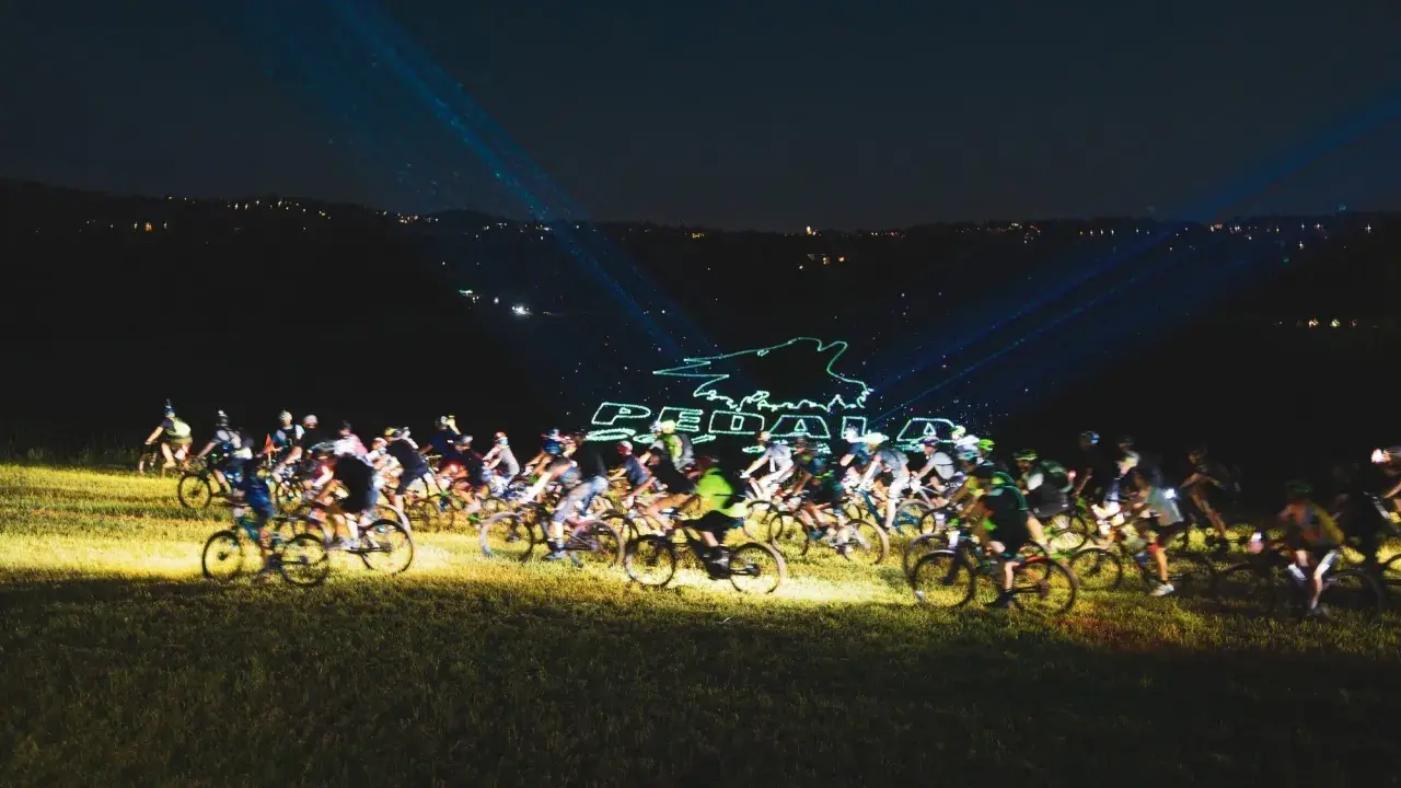 Ciclisti nella notte, illuminati da laser che formano il logo "Pedala coi Lupi". Un'avventura notturna su due ruote.