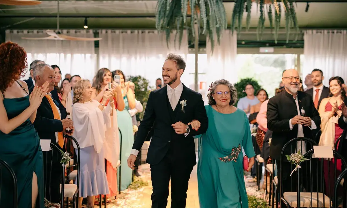 El novio sonriente camina del brazo de su madrina, ambos sonríen a los invitados.