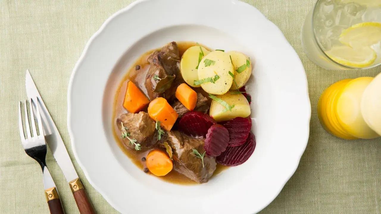 En tallrik med skånsk kalops, mört kött, morötter, potatis och rödbetor. Serveras med en drink och bestick.
