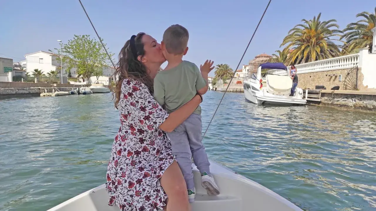 Madre besa a su hijo en un paseo en barco por los canales de Empuriabrava, con palmeras y casas blancas al fondo.