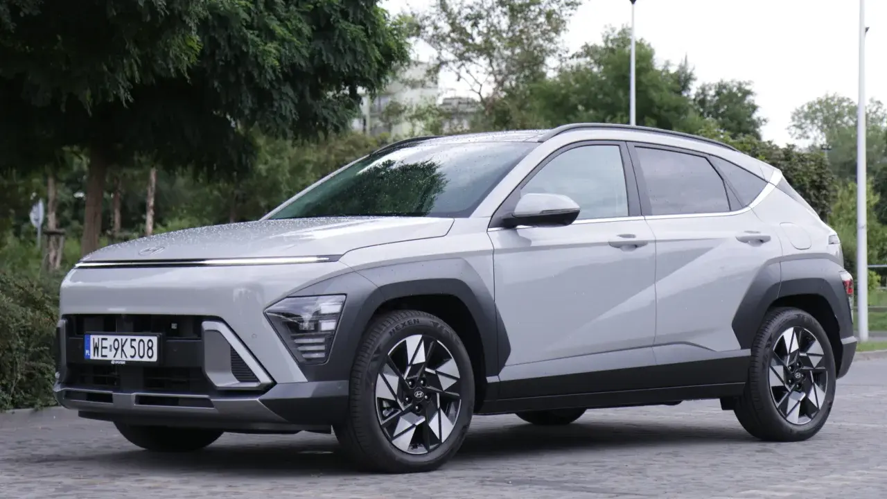 Nowy Hyundai Kona Hybrid w kolorze jasnoszarym, zaparkowany na brukowanej nawierzchni, otoczony zielenią.