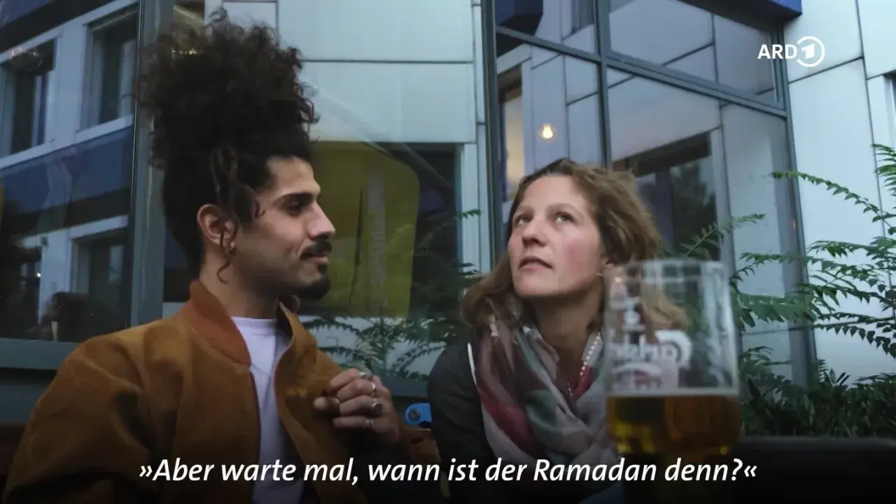 Ein Mann mit vollem Haar und eine Frau sprechen. Er fragt: "Aber warte mal, wann ist der Ramadan denn?" Die Fastenzeit im Islam ist ein wichtiges Thema.