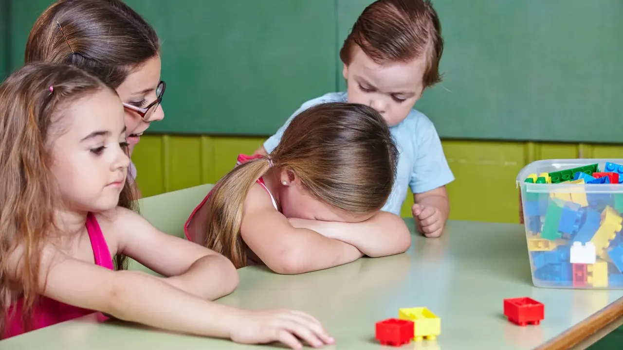 Smutna dziewczynka z płaczem ukrywa twarz na stole, podczas gdy inne dzieci i nauczycielka próbują ją pocieszyć. To obraz nieudanej adaptacji w przedszkolu.