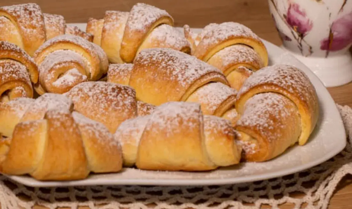 Talerz pełen puszystych, posypanych cukrem pudrem rogalików krucho drożdżowych. Idealne na śniadanie lub deser.