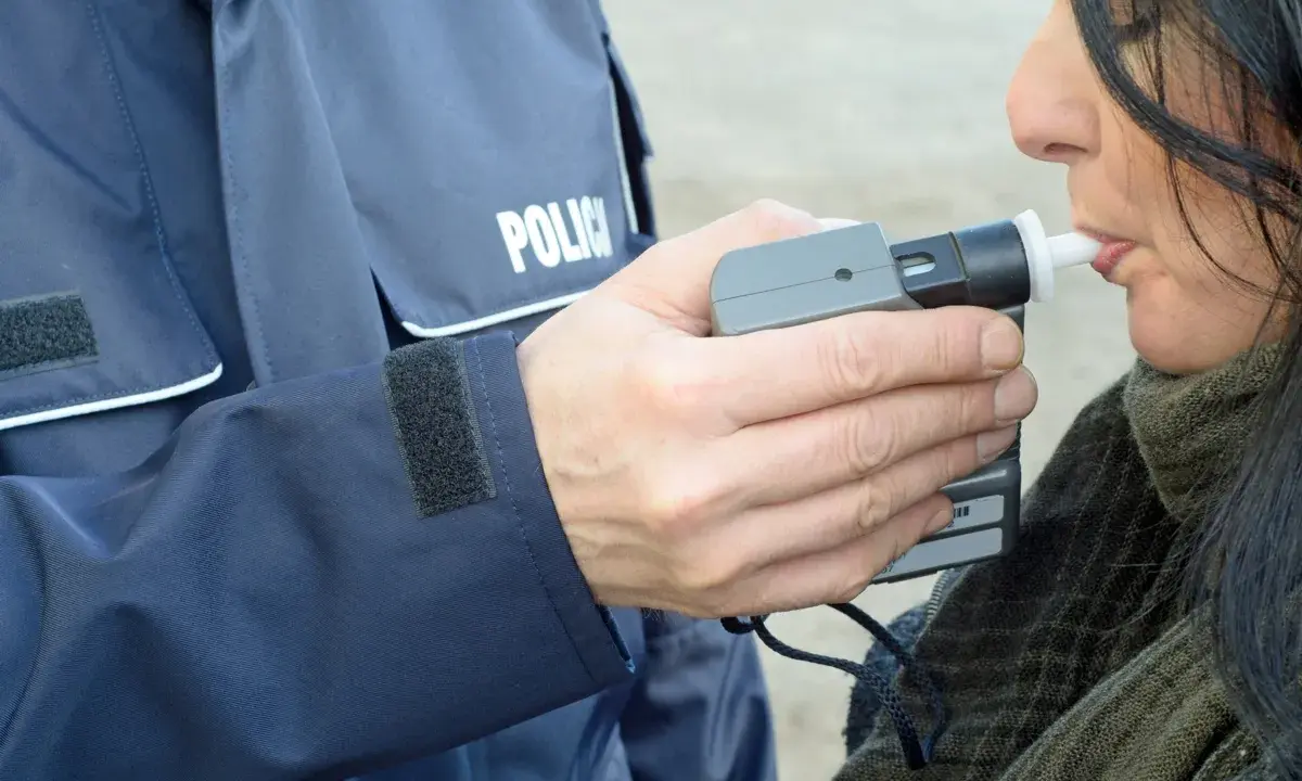Policjant w mundurze z napisem "POLICJA" mierzy kobiecie poziom alkoholu alkomatem.