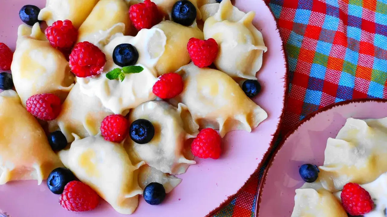 Pyszne pierogi z malinami i borówkami, polane śmietaną, na różowym talerzu. Letnia uczta pełna smaku.
