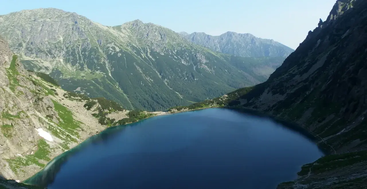 Górski krajobraz z jeziorem i stromymi zboczami. W oddali widać pasmo górskie, które przypomina najwyższy szczyt w Polsce.