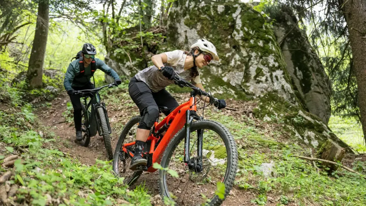 Dwie osoby na rowerach zjazdowych, jeden pomarańczowy rower bez amortyzatora z przodu, jadą po leśnym szlaku.