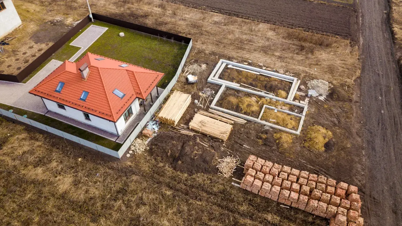Budowa domu na działce rolnej. Widać fundamenty, sterty drewna i cegieł, gotowy dom. To może być początek planowania, jak podzielić działkę rolną.
