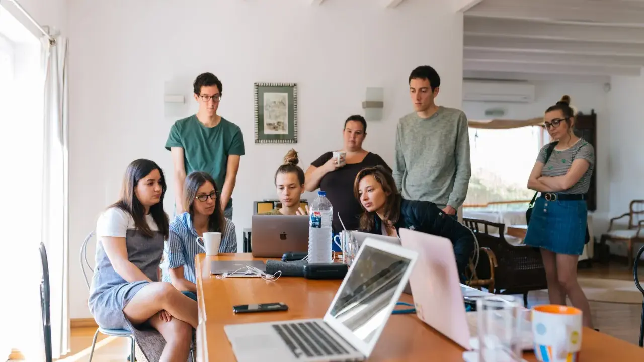 Grupa studentów na którym roku studiów są praktyki, pochylona nad laptopami, dyskutuje przy drewnianym stole.