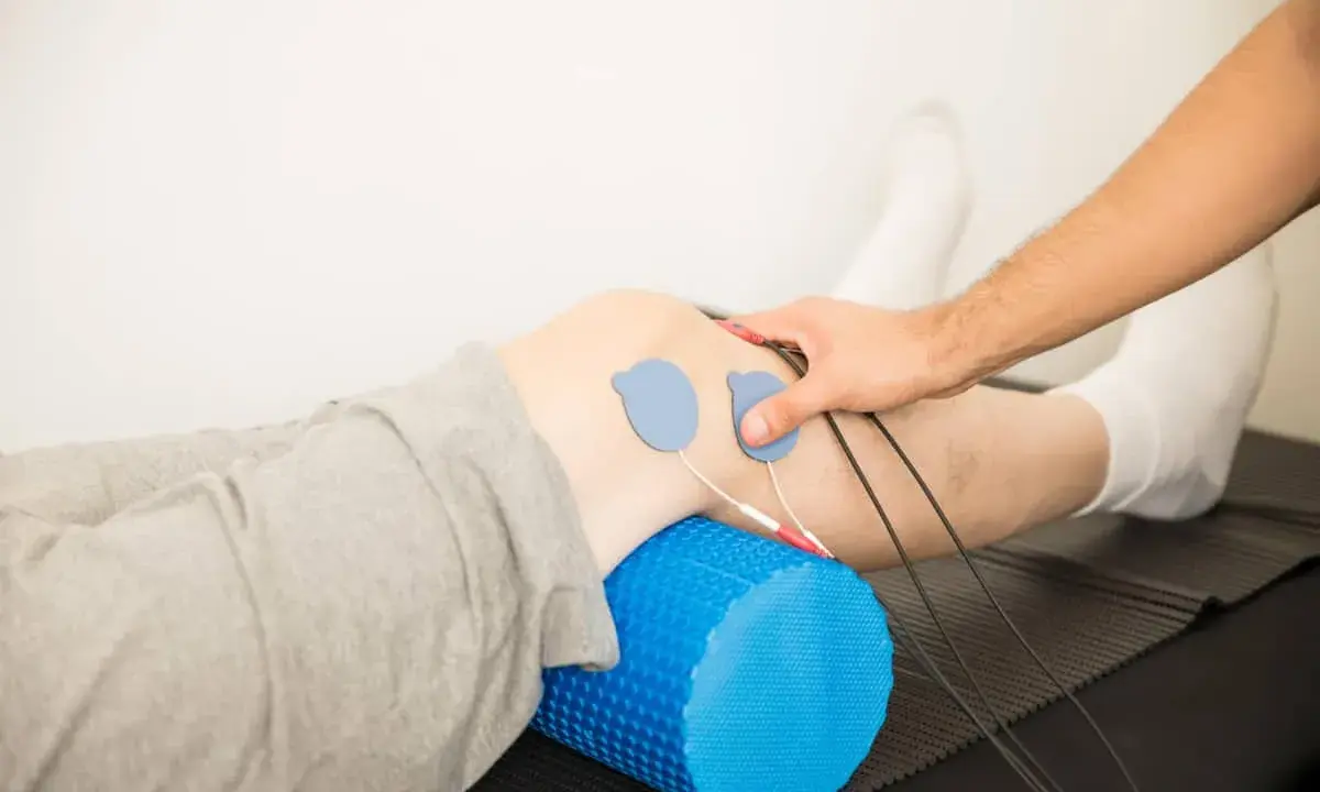 Rehabilitacja kolana w domu: elektrody na skórze, wałek do masażu pod nogą, ręka terapeuty.