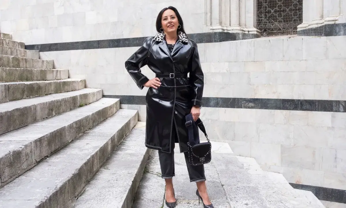 Donna con un audace outfit rock: trench in ecopelle nera, jeans scuri, décolleté borchiate e borsa a tracolla.