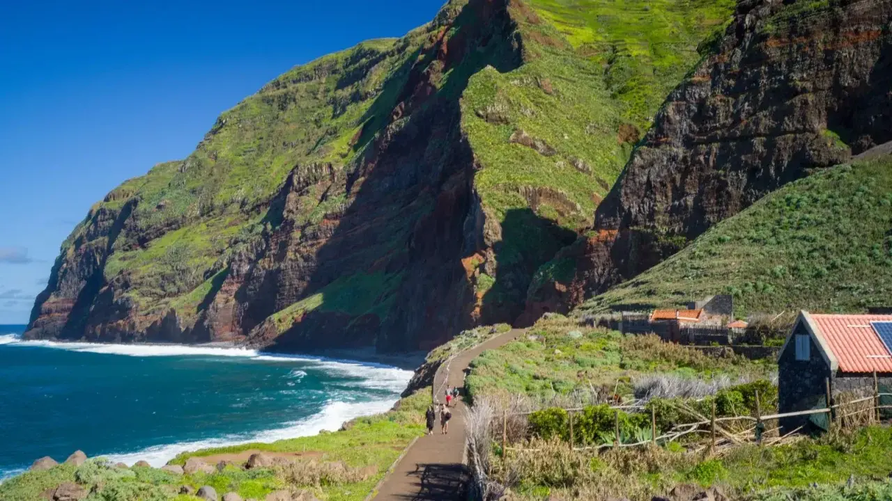 Grüne Berge und rote Felsen prägen die Küste der Madeira Inselgruppe. Ein Weg führt zu kleinen Häusern.