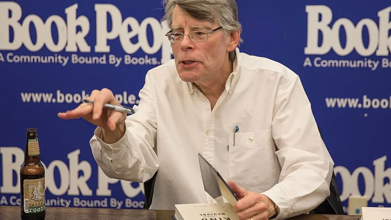 Stephen King, autore di libri da brivido, firma un volume. Sul tavolo, una bottiglia di root beer.