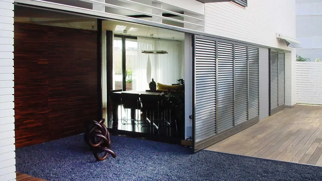 Una moderna casa con una contraventana corredera de lamas metálicas que protege un comedor interior.