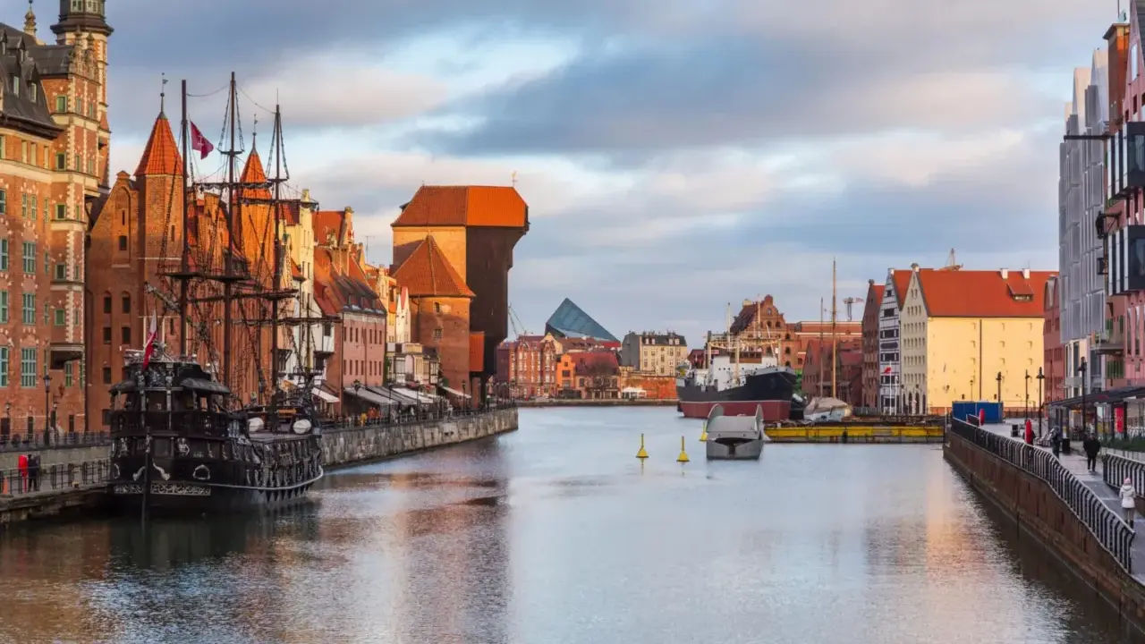 Gdańsk: zabytkowy żaglowiec cumuje przy Motławie, w tle Żuraw i nowoczesne budynki. Malowniczy opis miasta.