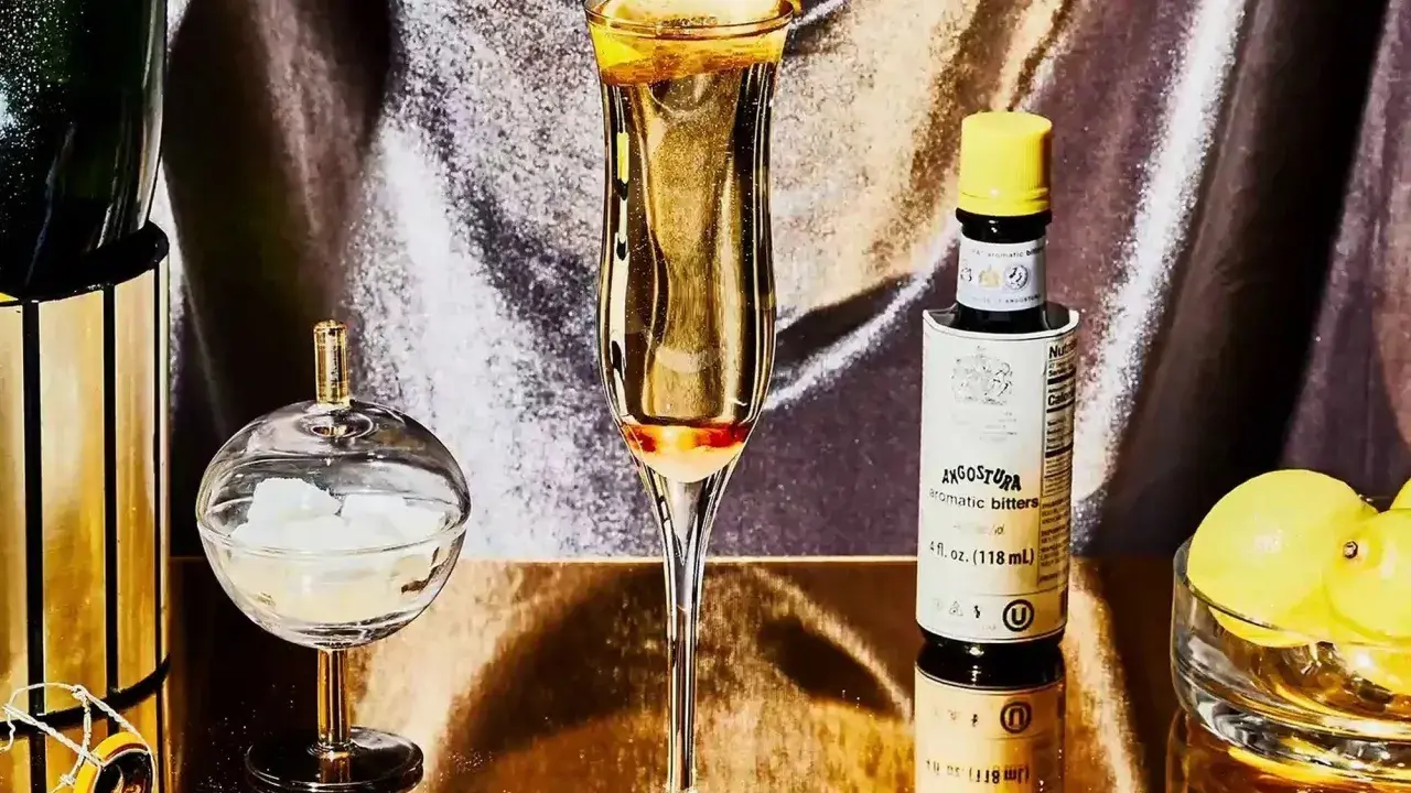 Elegantes Glas mit einem Champagner-Cocktail, garniert mit Zitronenschale. Daneben eine Flasche Angostura, Zuckerwürfel und Zitronen.