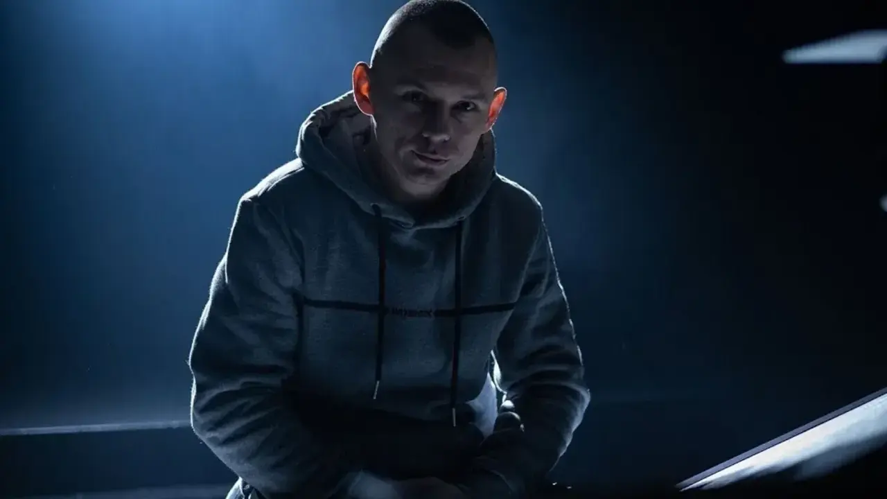 Młody mężczyzna w szarej bluzie z kapturem, z ogoloną głową, w mrocznym świetle. Jego spojrzenie sugeruje opowieść o polskim rapie.