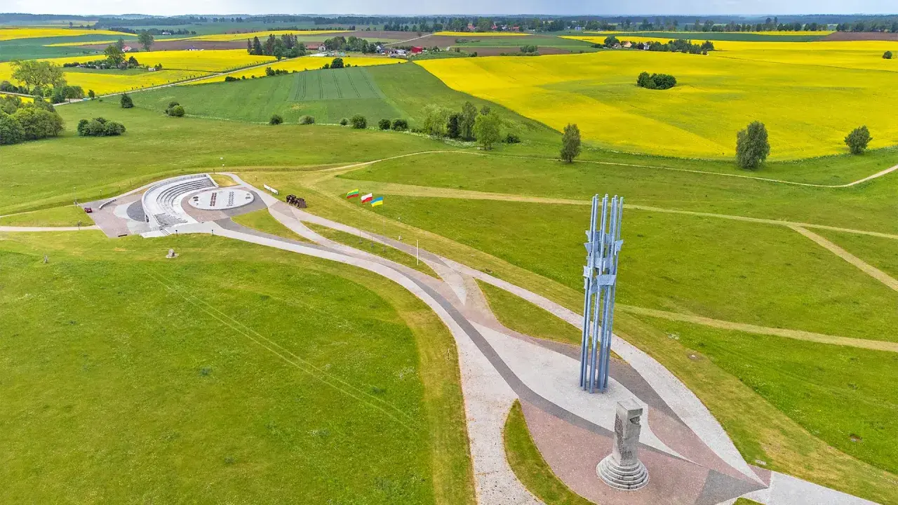 Pomnik i amfiteatr na polach grunwaldzkich, otoczone zielonymi łąkami i kwitnącymi polami rzepaku.