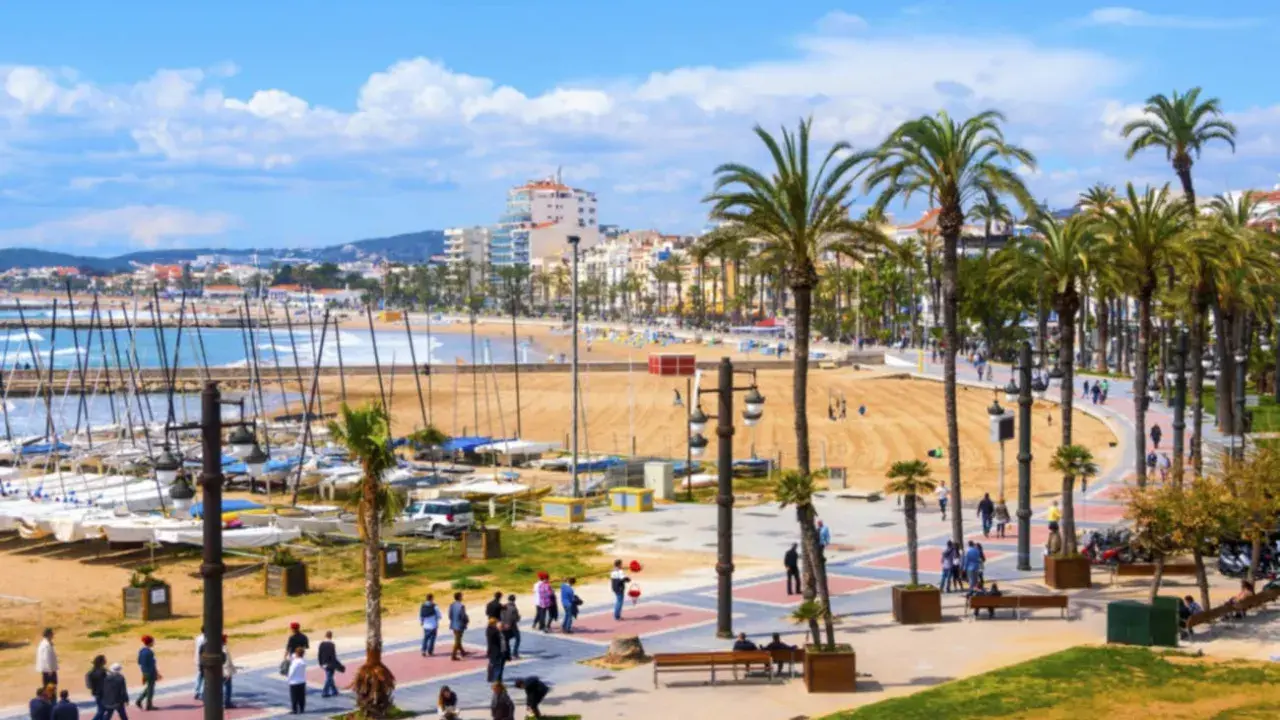 En livlig strandpromenad i Sitges, Spanien, med palmer, en sandstrand, båtar i hamnen och människor som njuter av solen.