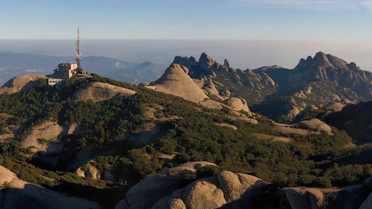 Edificio con antena en la cima de una montaña rocosa, con vistas a un paisaje montañoso y brumoso que se extiende hasta el horizonte, evocando la sensación de estar en planos cerca de Barcelona.