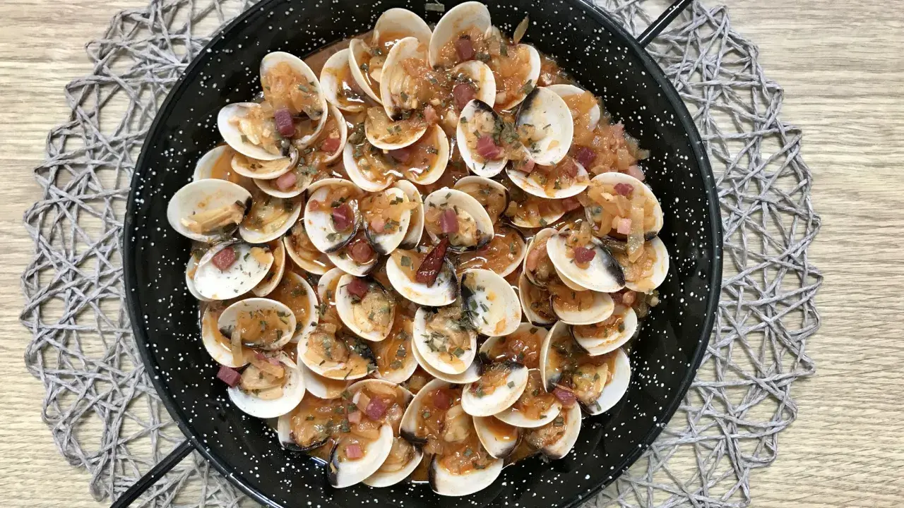 Almejas con jamón en una paellera, con trocitos de jamón y perejil picado, sobre un mantel de madera.
