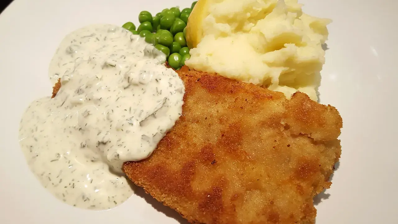 Panerad fisk med krämig kall sås till, serverad med potatismos, gröna ärtor och en citronklyfta.