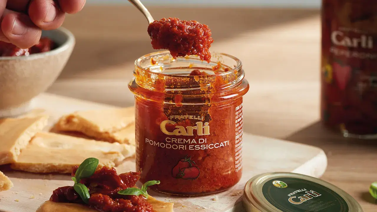 Une cuillère prélève de la crème de tomates séchées d'un pot Carli. Servie sur des crackers avec du basilic.