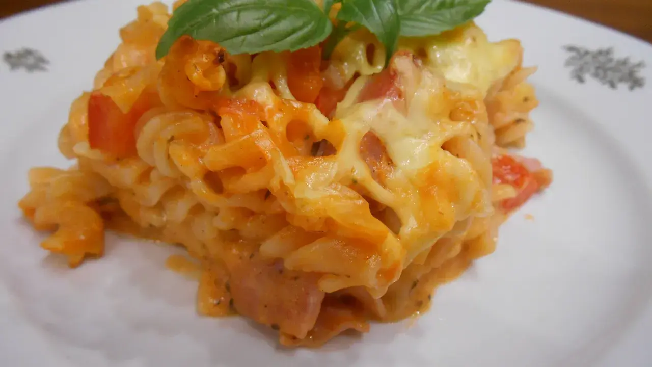 Pyszna zapiekanka z szynką, makaronem fusilli, pomidorami i ciągnącym się serem, ozdobiona listkami bazylii.