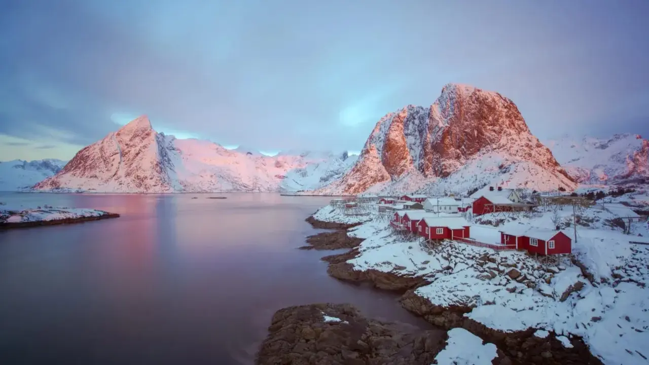 Vackert lofoten vinterlandskap med snöklädda berg, röda hus vid vattnet och en rosa himmel.