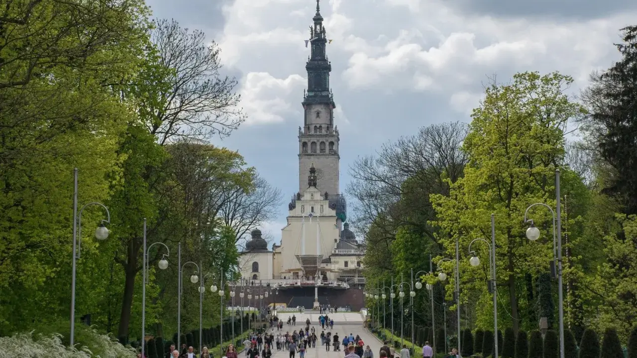 Aleja prowadząca do imponującej wieży klasztornej, jednego z ważnych sanktuariów w Polsce. Ludzie spacerują, podziwiając architekturę i otaczającą zieleń.