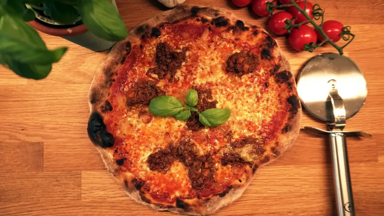 En napolitansk pizza med köttfärs som topping, garnerad med färsk basilika. Bredvid ligger en pizzaskärare och körsbärstomater.