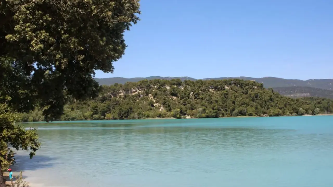 Vue paisible de l'étang de la Bonde, avec ses eaux turquoise bordées de verdure et de collines boisées sous un ciel bleu clair.