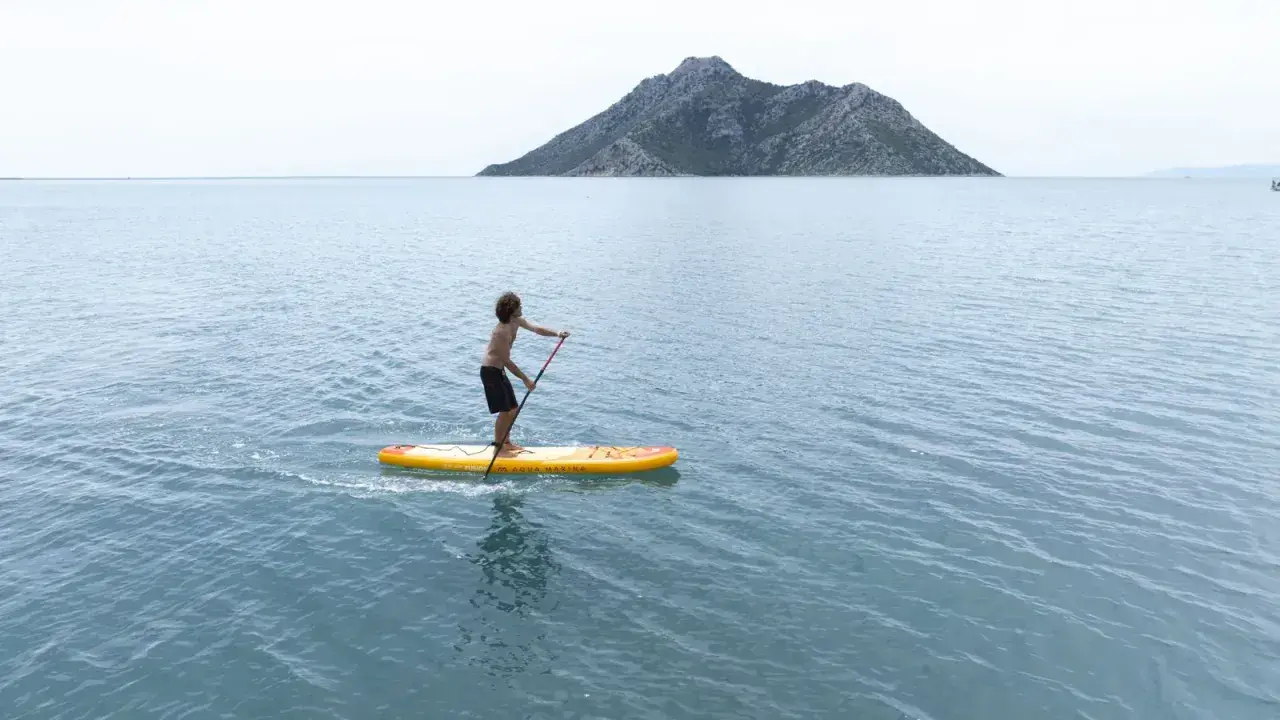 Chłopak na desce sup Aqua Marina przemierza spokojne morze w kierunku skalistej wyspy.