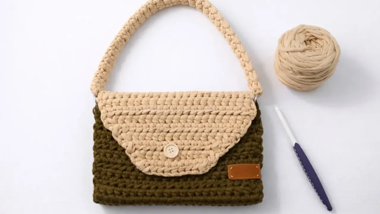 Bolso de trapillo bicolor, verde y beige, con asa y botón de madera. A su lado, ovillo de lana y ganchillo.