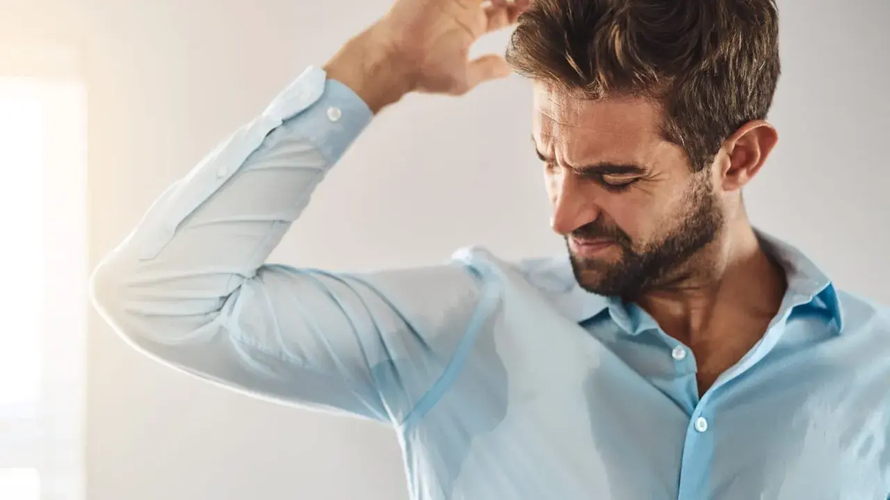 Uomo con camicia azzurra macchiata di sudore sotto le ascelle, che si gratta la testa. Sudare tanto è sintomo di stress o caldo.
